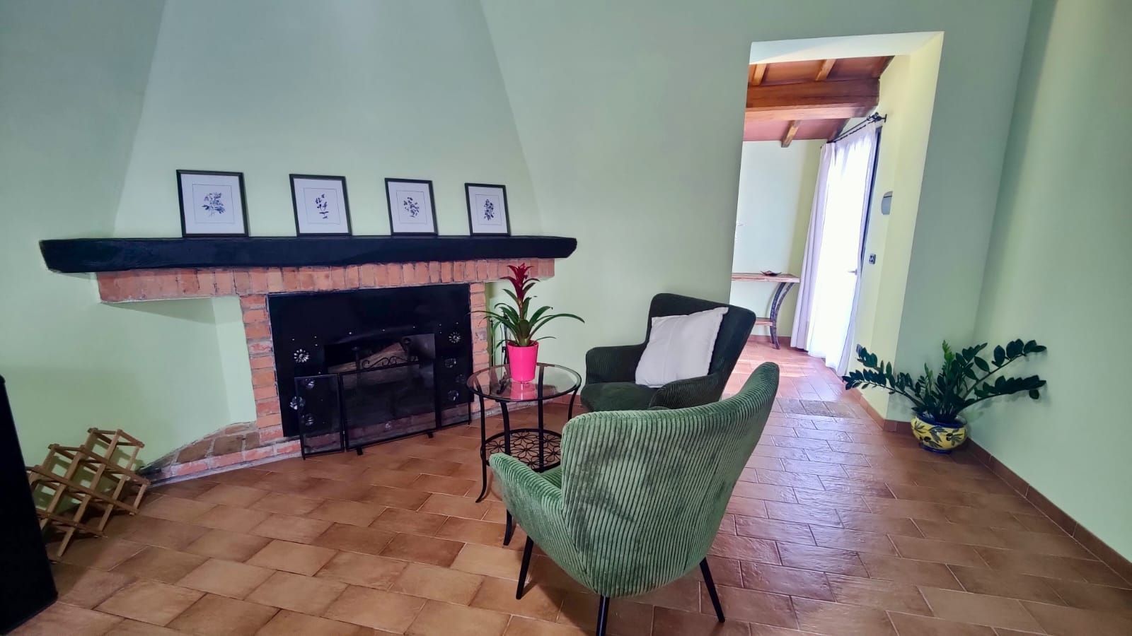 A cosy lounge area with mint green walls and a brick fireplace topped by four framed botanical prints. Two green upholstered chairs flank a small round glass table holding a potted plant with red flowers. To the right, a doorway opens into a sunlit room with a wooden ceiling and glass door. Several potted plants rest on the brown tiled floor near the fireplace.