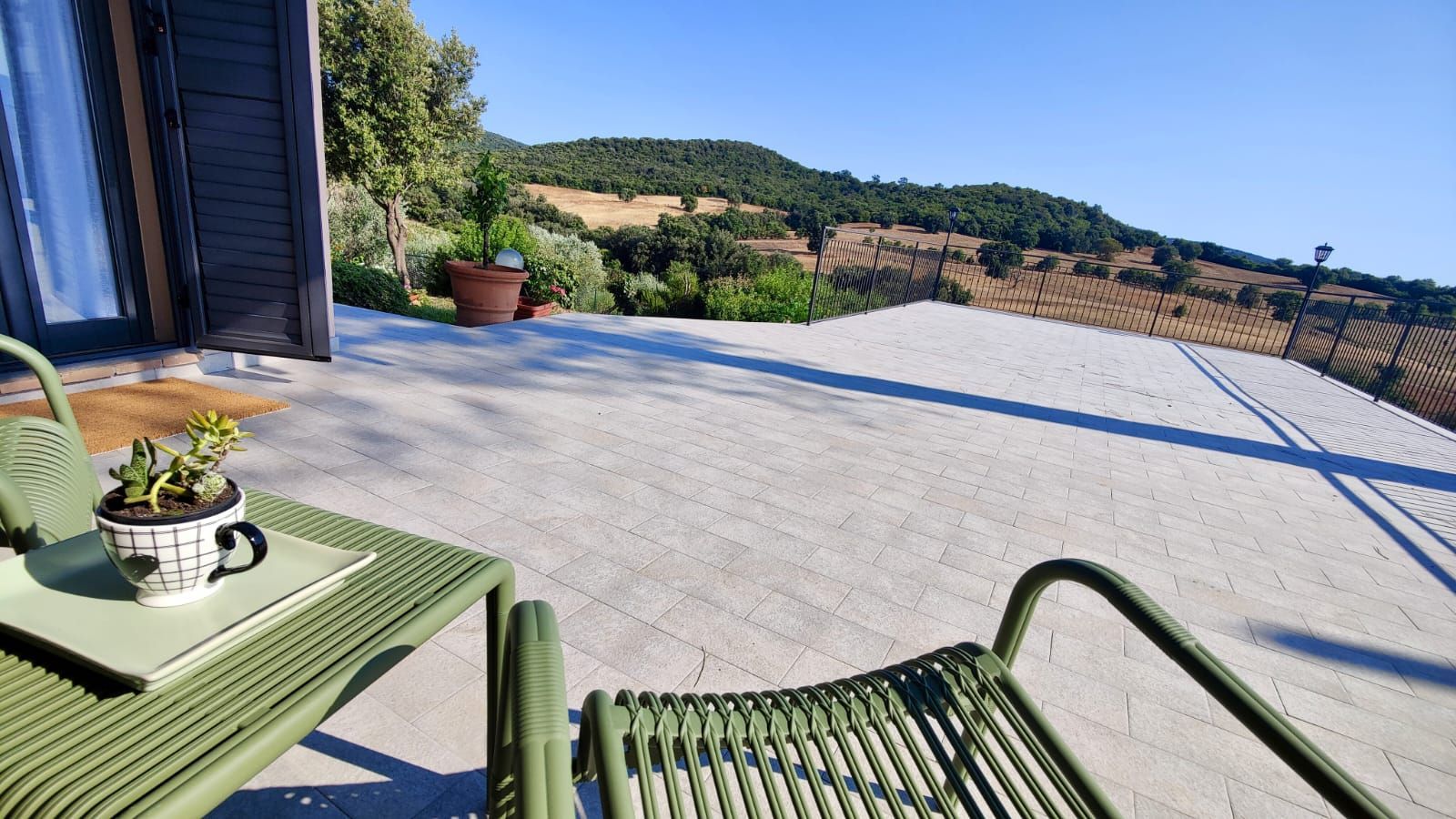 Two light-green chairs flank a small white table holding a potted succulent on a pale tiled patio bordered by a black metal railing, overlooking rolling green hills, trees, and fields beneath a clear blue sky.