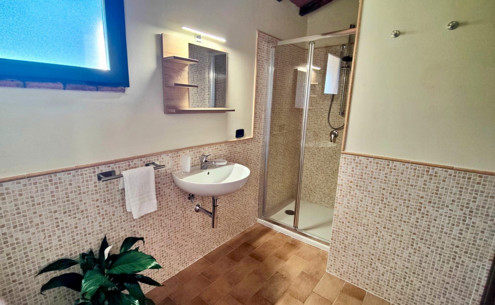 Modern minimalist bathroom with beige-and-white mosaic tile walls and brown floor tiles. A frosted blue-tinted window sits above a metal towel rack holding a white towel. Below, a wall-mounted white sink with chrome faucet is set beneath a wooden shelf and mirror with integrated lighting. To the right, a frameless glass shower features a chrome showerhead and hanging towel. A small green potted plant rests on the floor beside the sink.