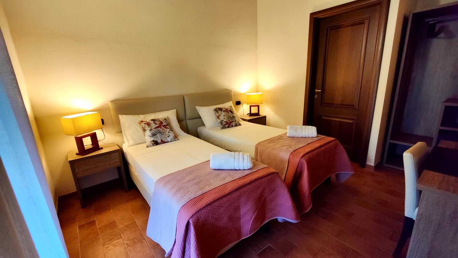 A cosy guest room featuring two single beds side by side. Each bed is dressed in a pink quilted coverlet, white linens, a folded white towel at the foot, and a floral-patterned pillow. Wooden nightstands with yellow-shaded lamps flank the beds. Behind them, a wooden door opens onto an open closet with built-in shelves. The terracotta-tiled floor leads to a simple desk and chair against the side wall.
