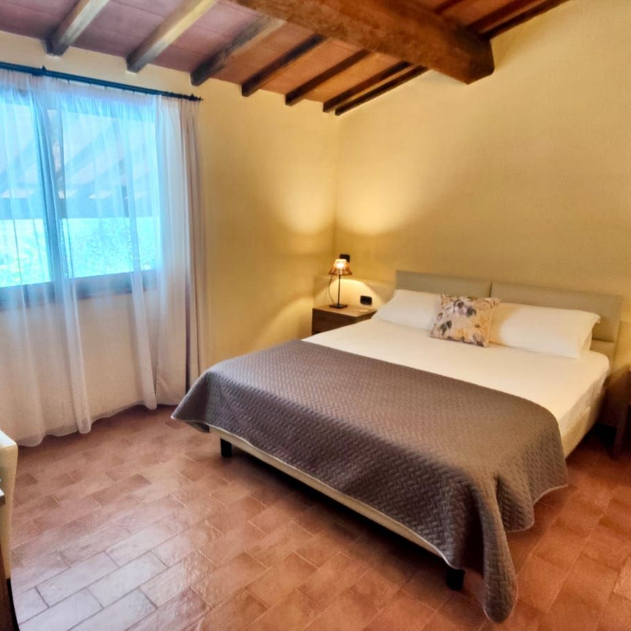 A cosy bedroom with a king bed dressed in a grey blanket and two pillows (one with a floral pattern). Exposed wooden beams form the sloped ceiling, and a large window with sheer curtains lets in soft light. Matching bedside tables hold lamps, and a small desk with a chair and a potted orchid sits against one wall. The floor is laid with warm terracotta tiles.