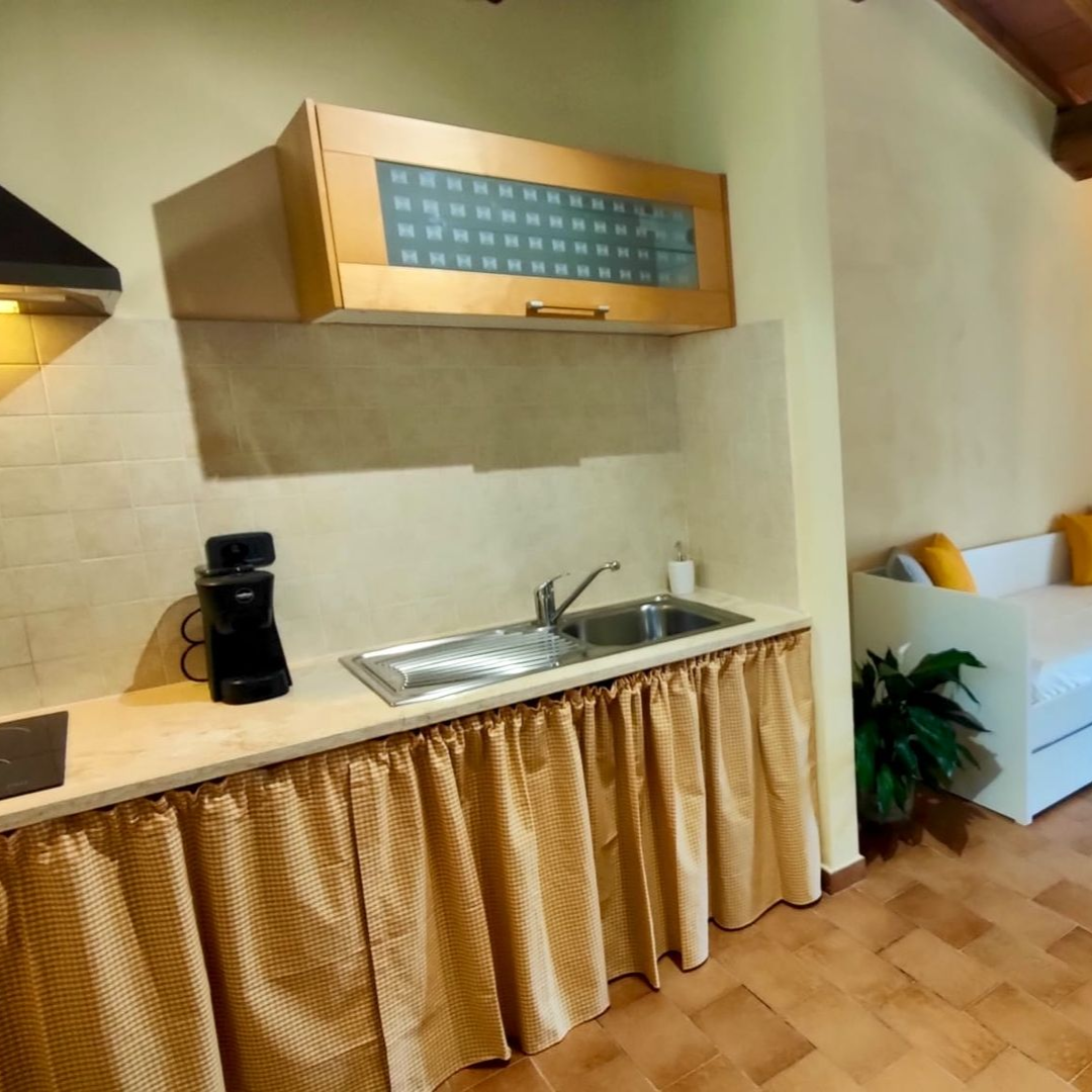 A cosy rustic kitchenette and living nook with exposed wooden ceiling beams and a tiled floor. A countertop holding a sink, stovetop and coffee maker is skirted by a green-and-white chequered curtain beneath frosted-glass cabinets. Beside it, a white daybed with yellow and grey cushions sits under sheer white curtains, with potted plants and a vase of white flowers adding a touch of greenery.