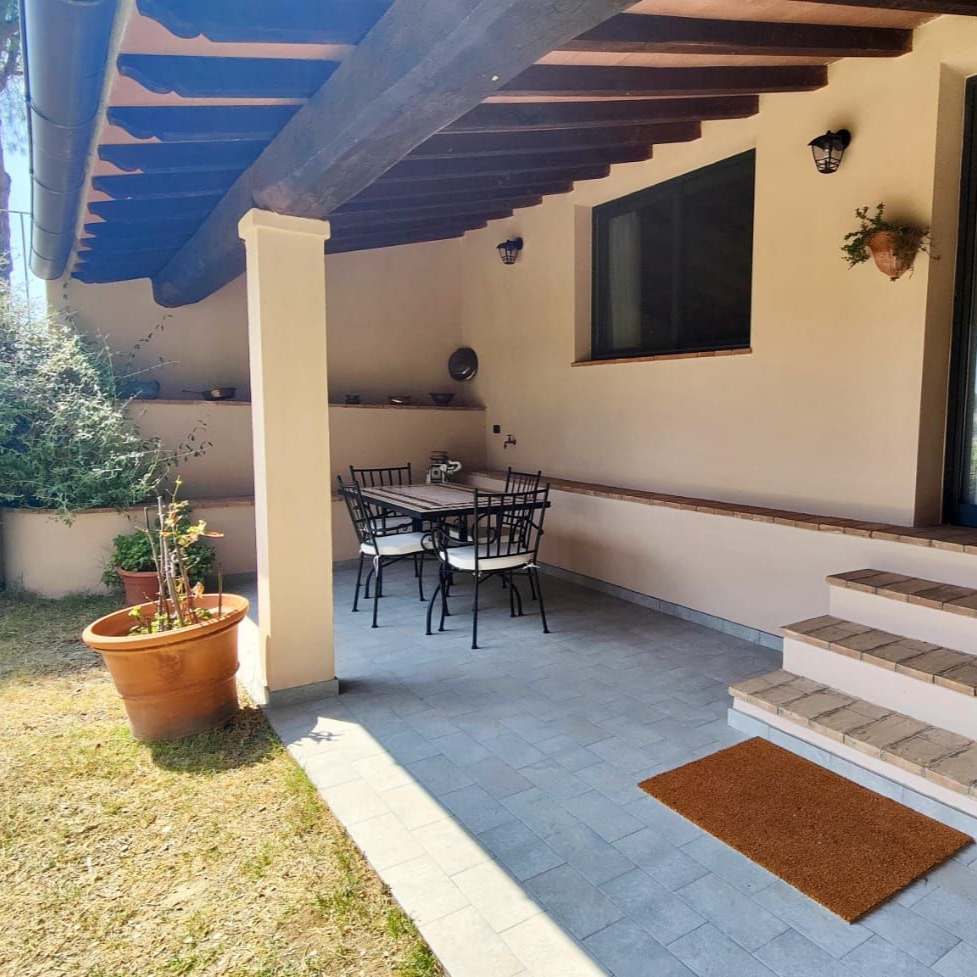 A tiled outdoor patio attached to an apartment, sheltered by a wooden roof on a single column. A small table with four chairs, a potted plant, and a doormat sit in front of large windows and a glass door. Beyond the patio, lush greenery and open fields stretch into the distance.
