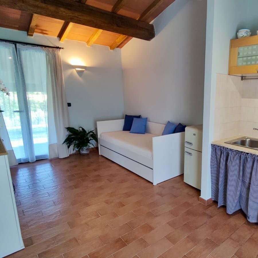 A cosy rustic room with exposed wooden ceiling beams. Along one wall, a small kitchenette features a sink, coffee maker, and stovetop hidden behind a blue-and-white chequered curtain, with glass-fronted wooden cabinets above. Opposite, a white loveseat topped with blue cushions sits beside a compact white refrigerator under a large window dressed in sheer curtains. A small table holding a potted plant and a floral arrangement adds a touch of greenery.