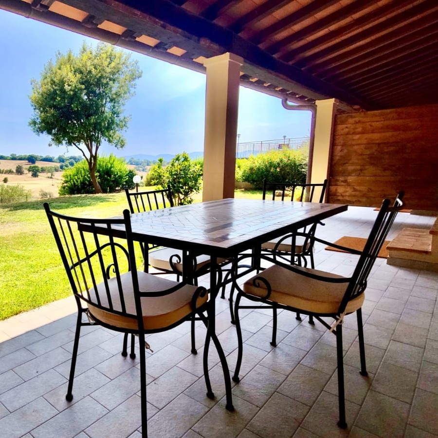 Covered outdoor patio with tiled floor and exposed wooden beam ceiling. A rectangular metal table is surrounded by cushioned chairs. Beyond the patio, a lush green lawn edged with shrubs and trees opens onto rolling hills under a clear, sunlit sky.