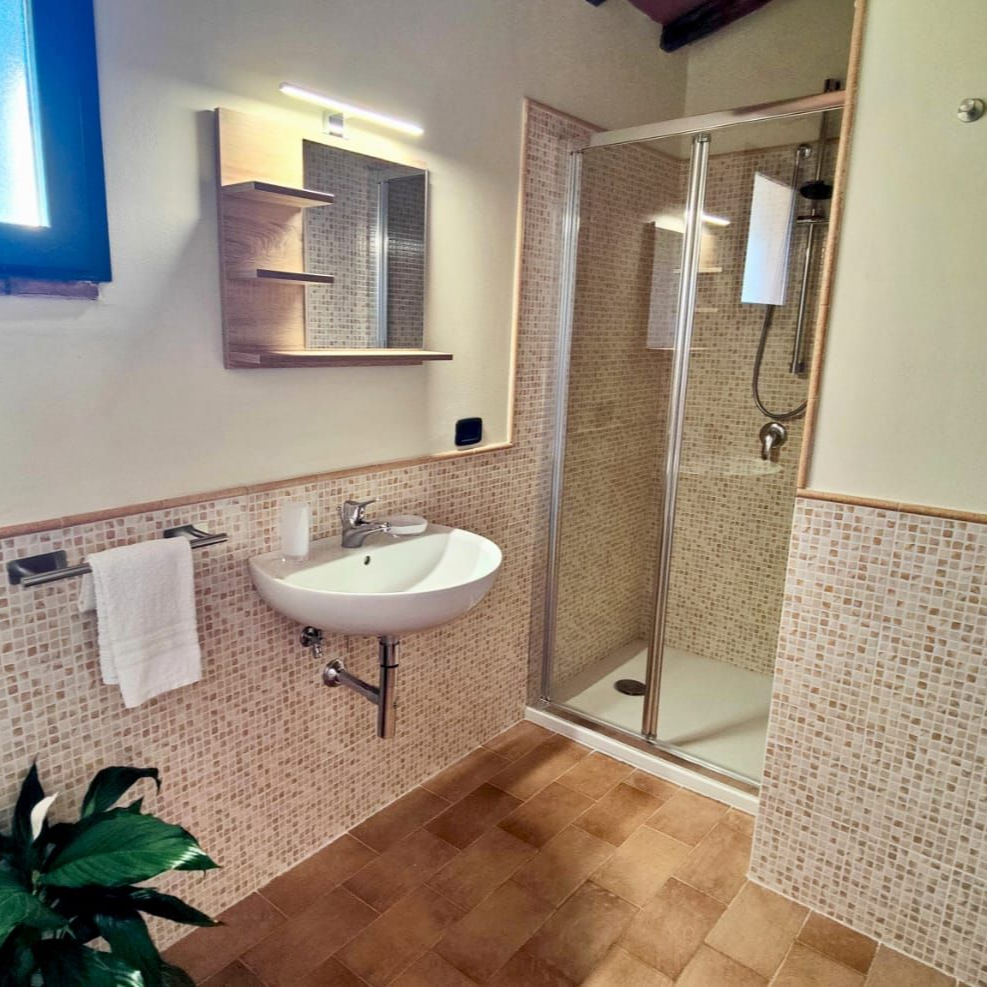 Modern minimalist bathroom with beige-and-white mosaic tile walls and brown floor tiles. A frosted blue-tinted window sits above a metal towel rack holding a white towel. Below, a wall-mounted white sink with chrome faucet is set beneath a wooden shelf and mirror with integrated lighting. To the right, a frameless glass shower features a chrome showerhead and hanging towel. A small green potted plant rests on the floor beside the sink.