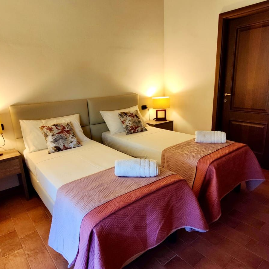 A cosy guest room featuring two single beds side by side. Each bed is dressed in a pink quilted coverlet, white linens, a folded white towel at the foot, and a floral-patterned pillow. Wooden nightstands with yellow-shaded lamps flank the beds. Behind them, a wooden door opens onto an open closet with built-in shelves. The terracotta-tiled floor leads to a simple desk and chair against the side wall.
