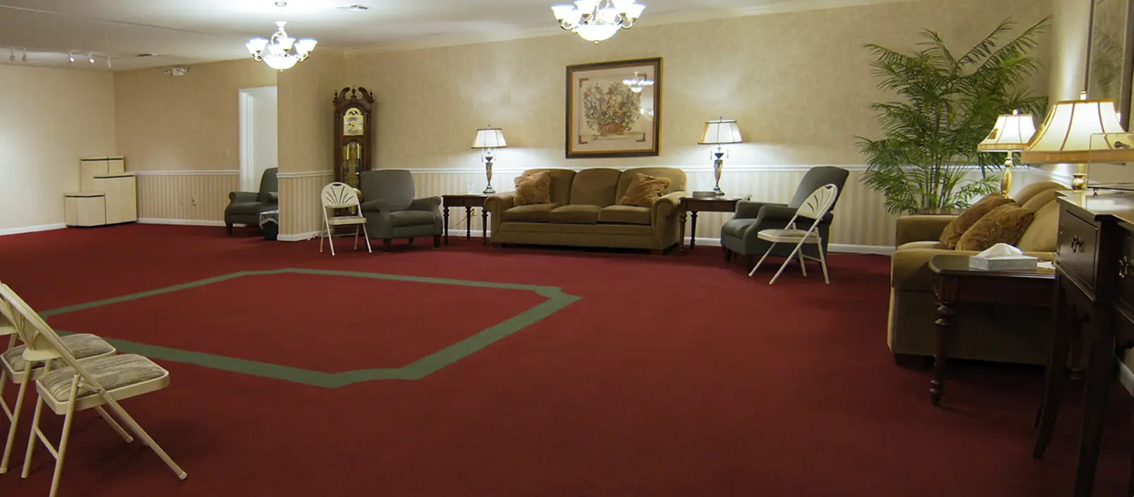 A warmly lit room with red carpet, sofas, chairs, a grandfather clock, and a plant.