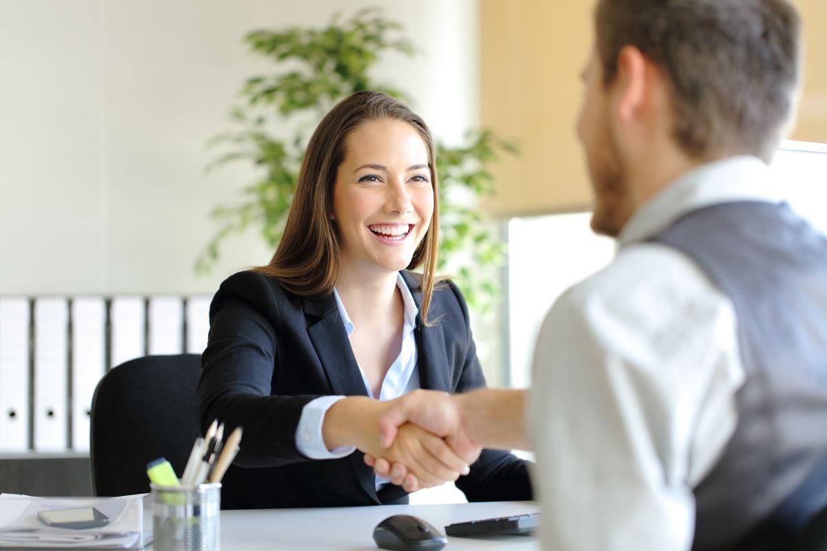 Woman in a black suit smiles, shaking hands with a man in an office setting.