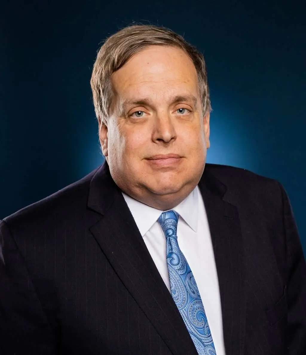 Man in suit and tie, looking at the viewer against a blue background.