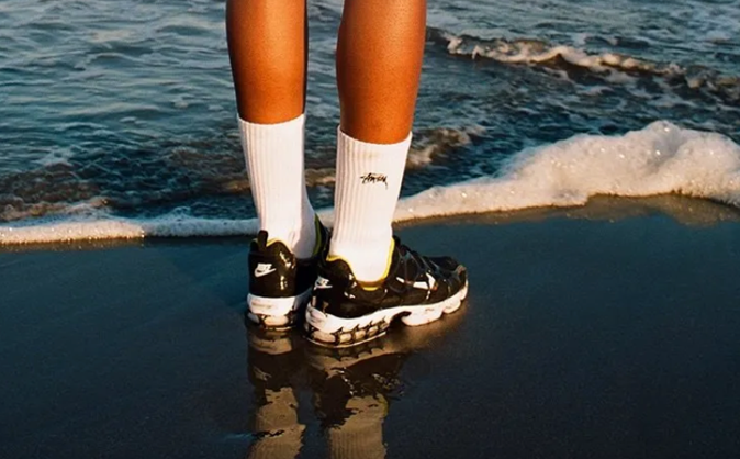 Image of beach front water with persons feet and legs wearing Nike shoes and socks