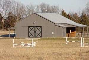 Gray barn with black cross-buck doors, small porch, and horse jumps in a grassy field.