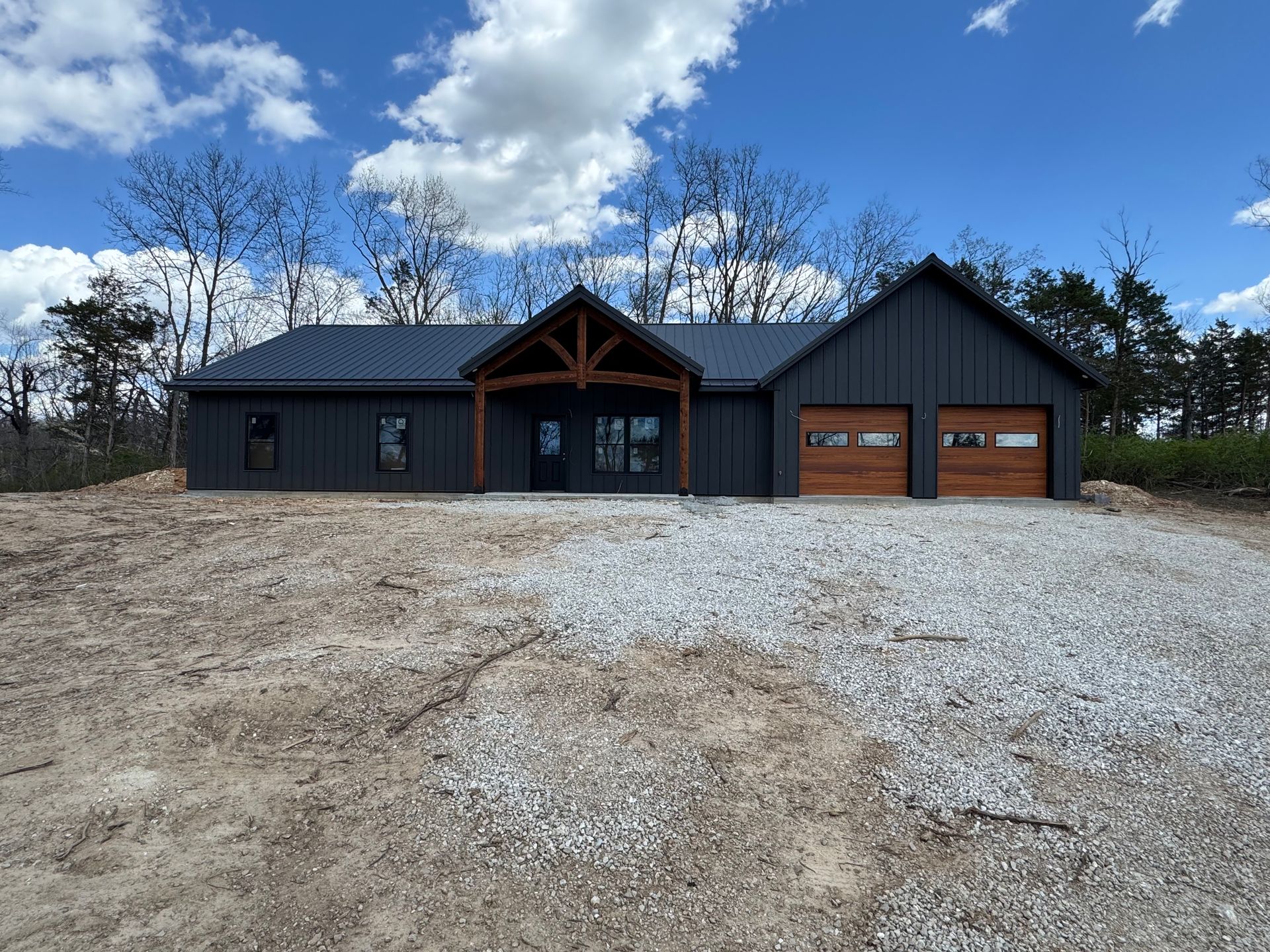 Dark gray modern home with brown garage doors and timber framing on a gravel lot under a cloudy blue sky.