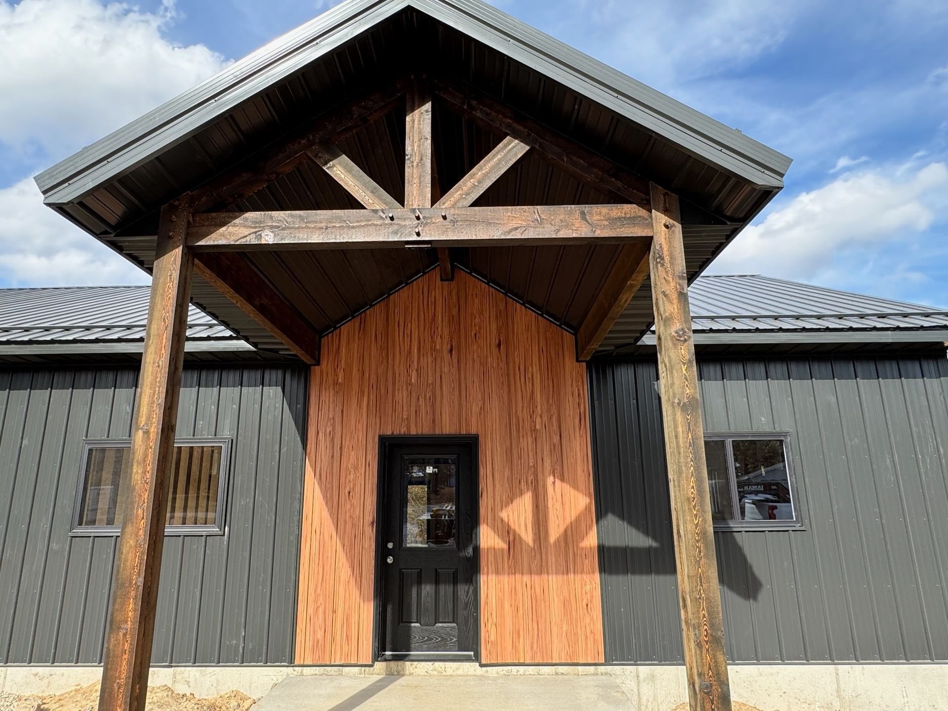 Entrance with wooden posts, gray siding, black door and roof, and a reddish-brown wood accent.