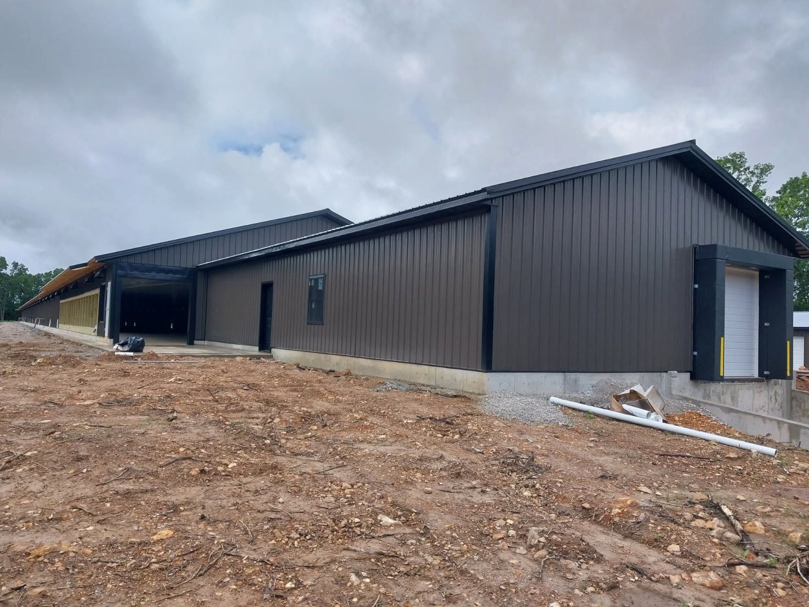 Brown metal building under construction, set on a dirt hill with a loading dock. Overcast sky.