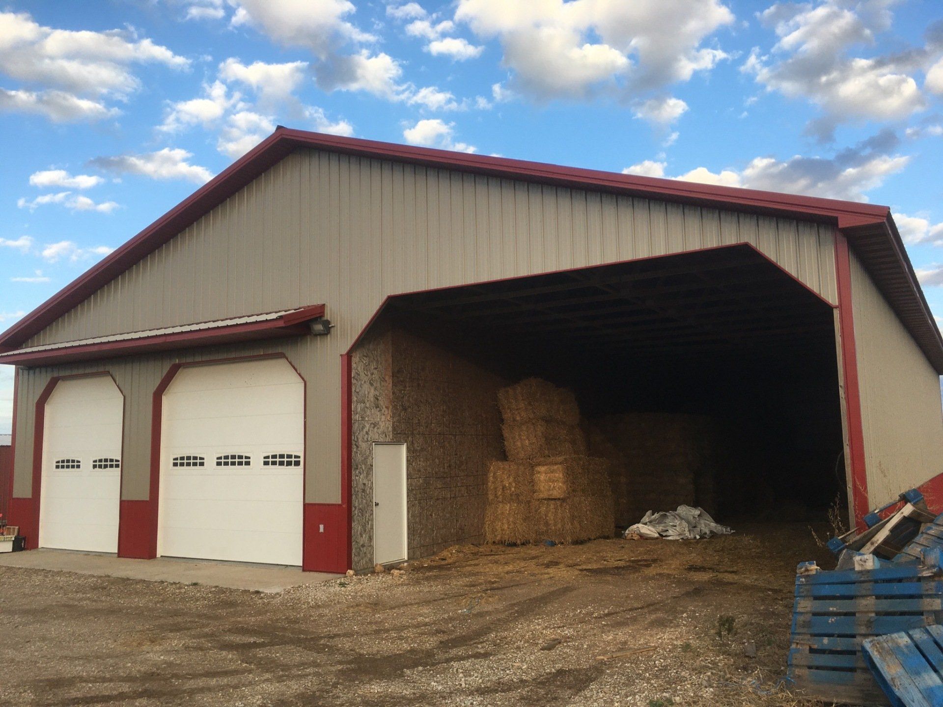 Barn with two white garage doors and an open bay filled with hay bales. Red trim and roof.