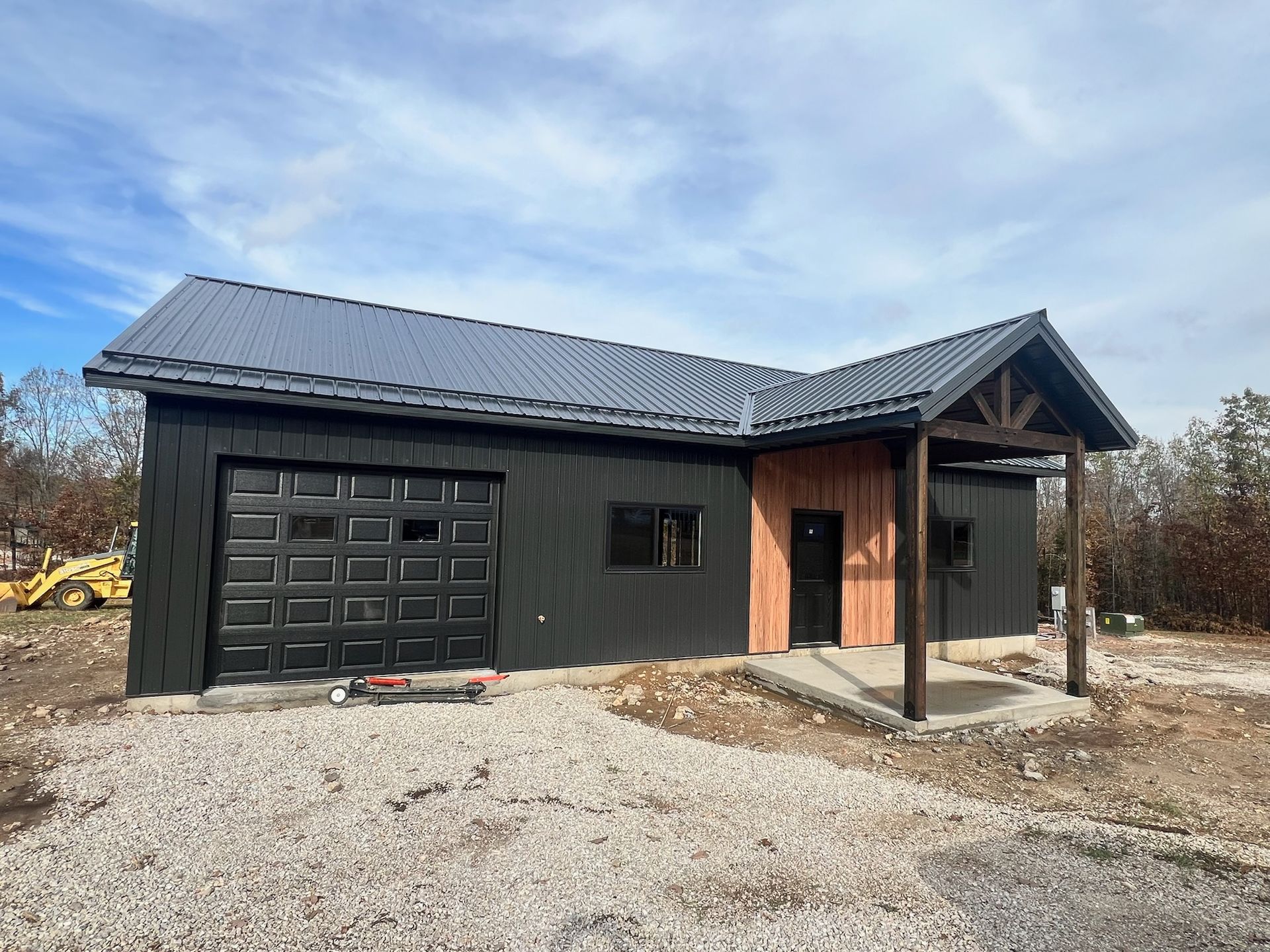 Black and brown modern building with a garage door, porch, and gravel yard under a cloudy sky.