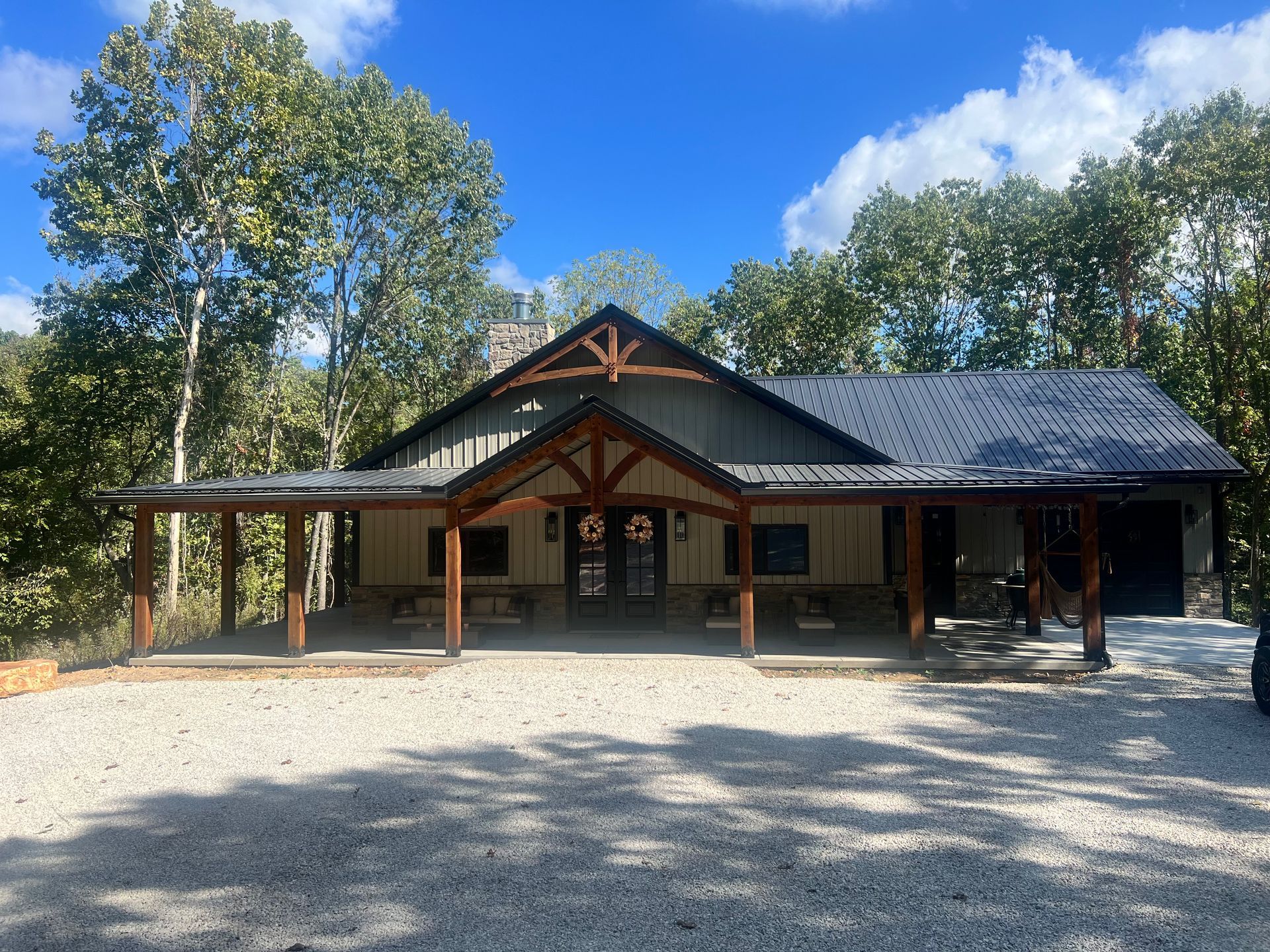 Cabin with covered porch and gravel driveway, surrounded by trees, under blue sky.