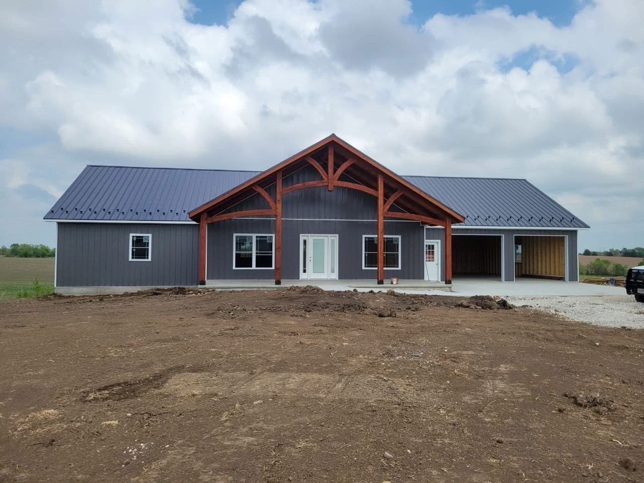 Gray house with a wooden portico and attached garage, set in a field under a cloudy sky.