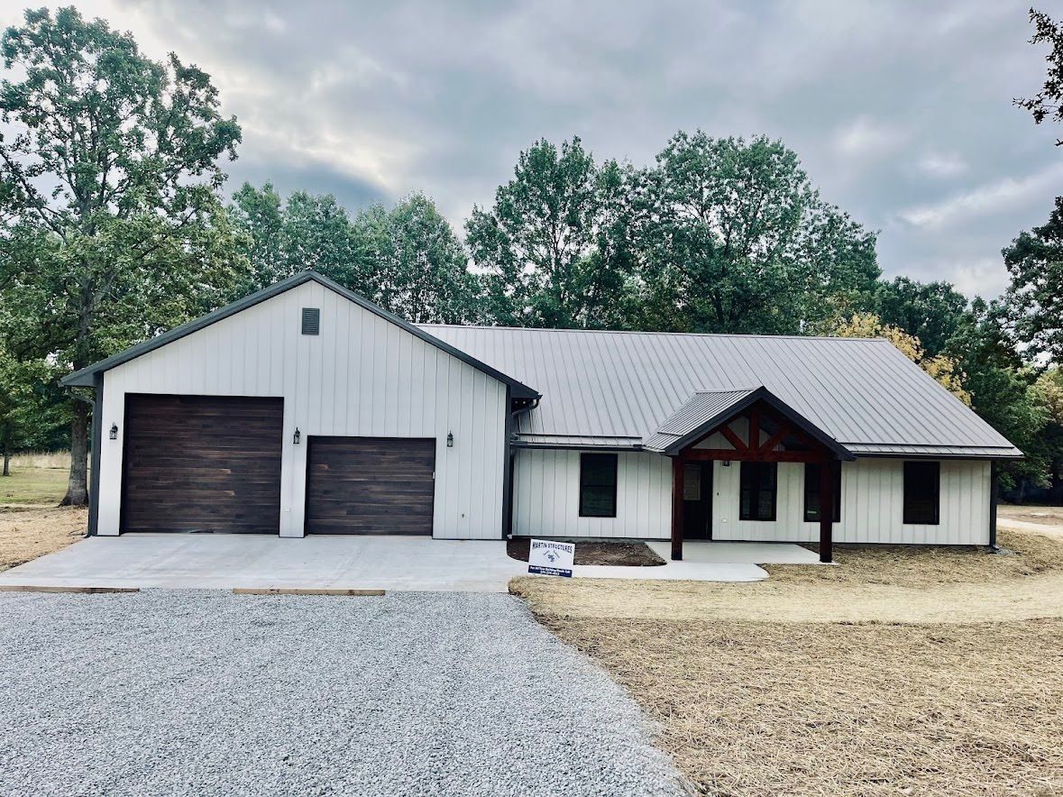 White farmhouse with gray metal roof and brown garage doors; gravel driveway.