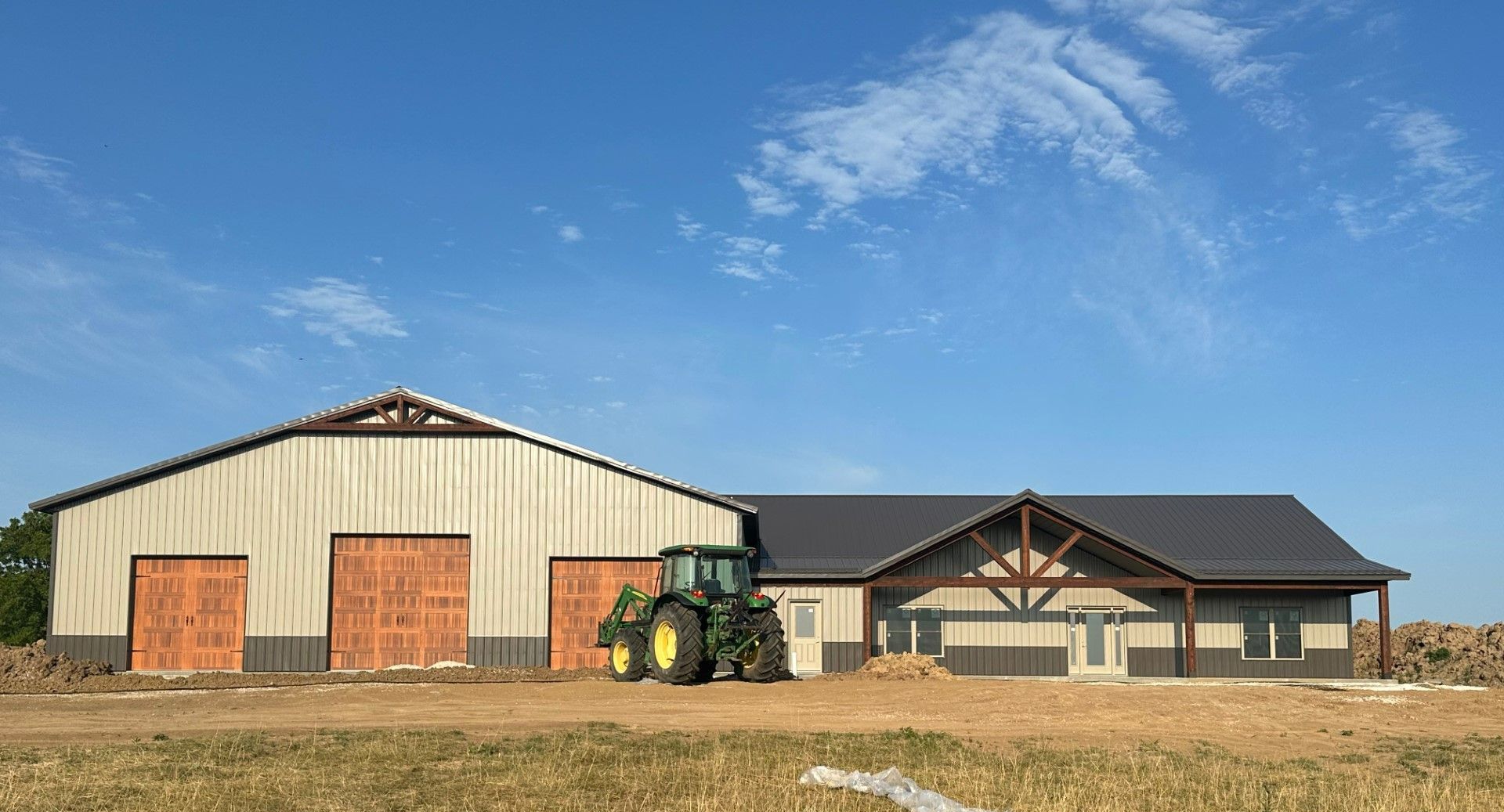 Barn and building under construction with John Deere tractor; tan and brown tones against blue sky.
