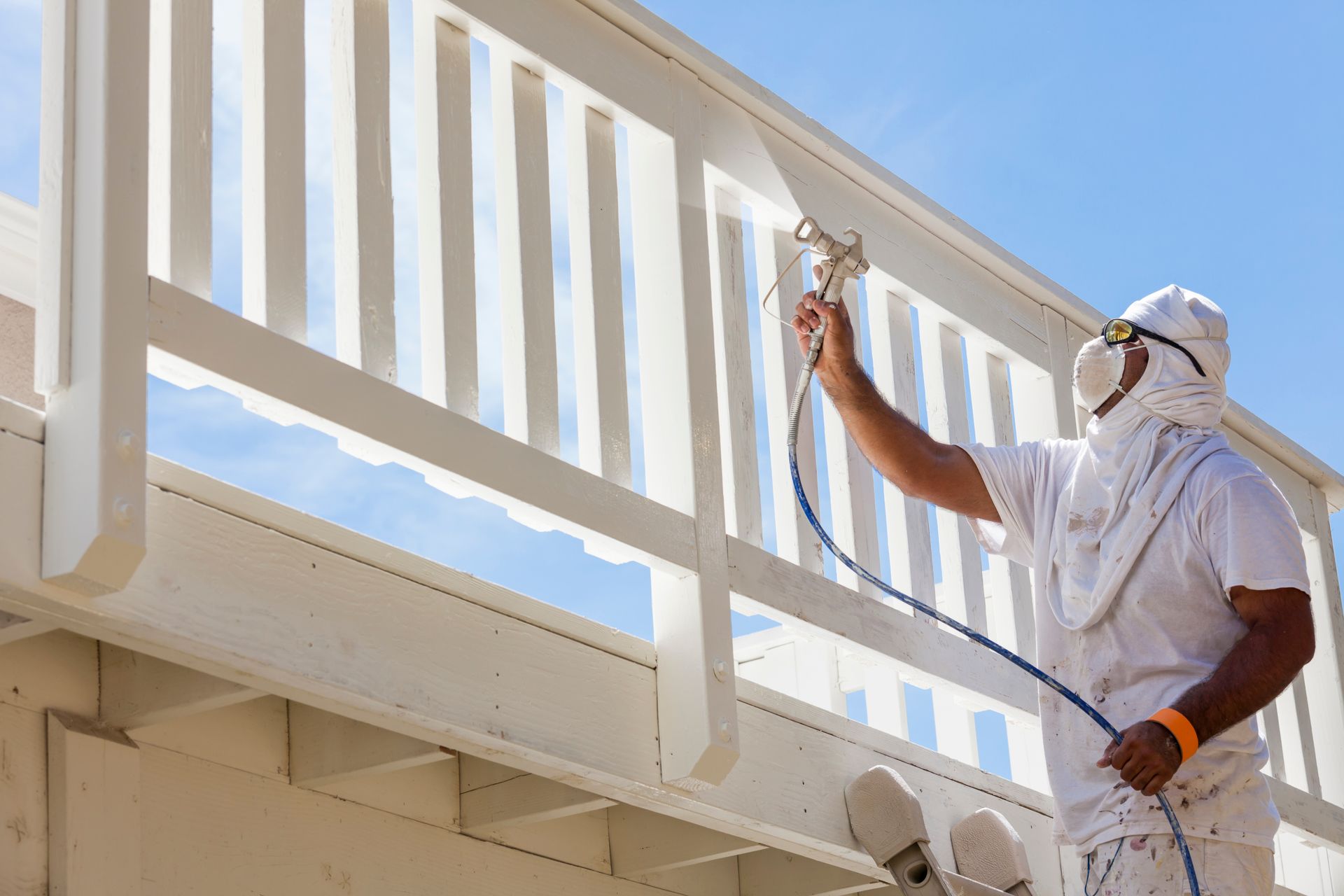 Person in white protective gear sprays white paint on a wooden railing outdoors.