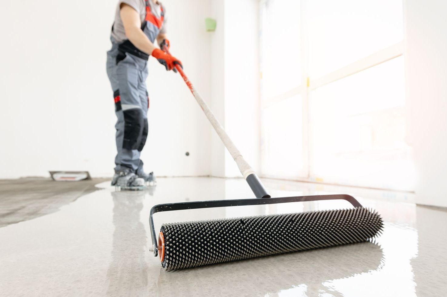 Person using a spiked roller on a shiny, light-colored floor; indoor, construction setting.