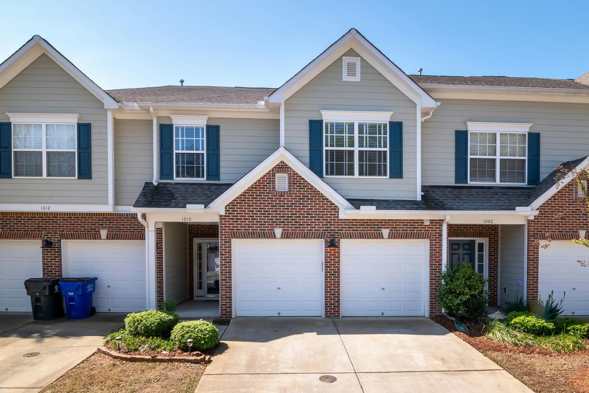 Townhouses with beige siding, brick accents, and white garage doors under a clear blue sky.