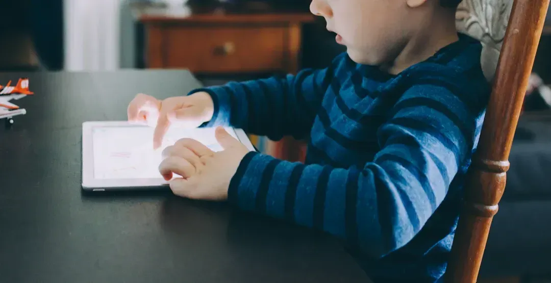 Child using a tablet, seated at a dark table. Blue striped shirt, indoor setting.