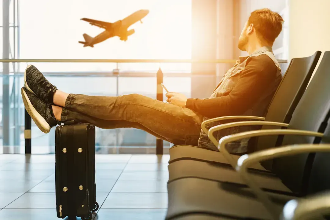Man at airport with feet up, looking at a tablet while plane takes off. Luggage by chair.