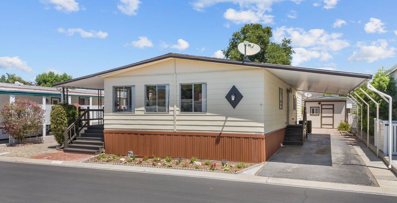 A beige mobile home with brown skirting, a front porch, and an attached carport on a paved street under a blue sky.