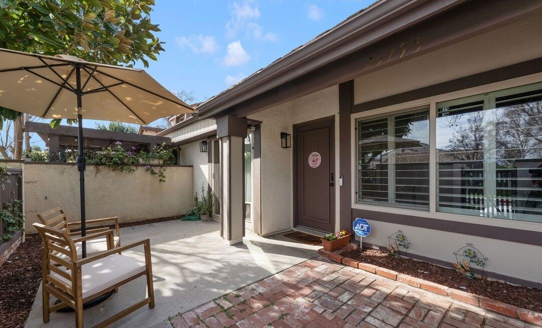 A patio area with two wooden chairs under a beige umbrella next to a house with a brick walkway and large window.