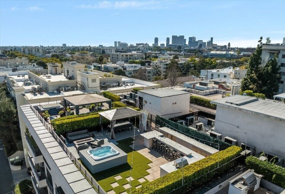 An elevated view of a modern rooftop terrace featuring a hot tub, lounge seating, dining areas, and a city skyline backdrop.