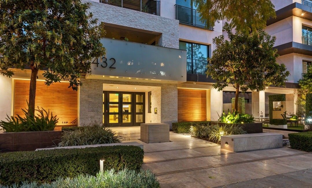The entrance to a modern multi-story building at dusk, featuring stone walls, warm lighting, and manicured landscaping.