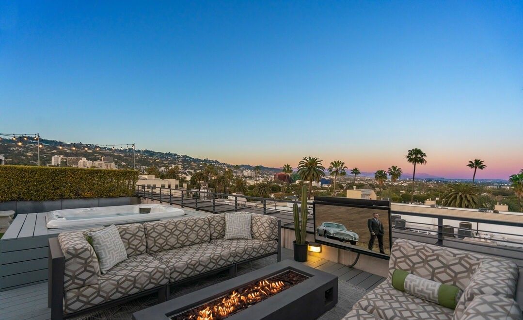 A rooftop terrace at sunset featuring a fireplace, patterned outdoor seating, a hot tub, and a large movie screen.
