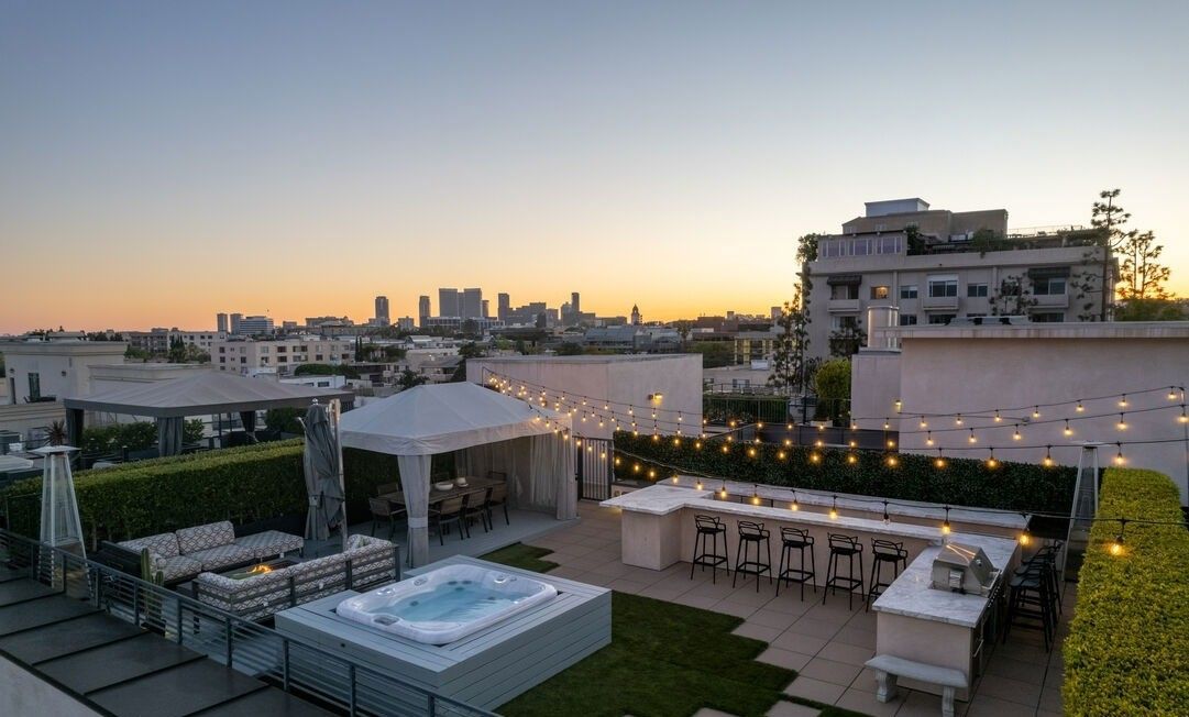 A luxury rooftop terrace at sunset featuring a hot tub, lounge seating, a bar, and a view of a city skyline.