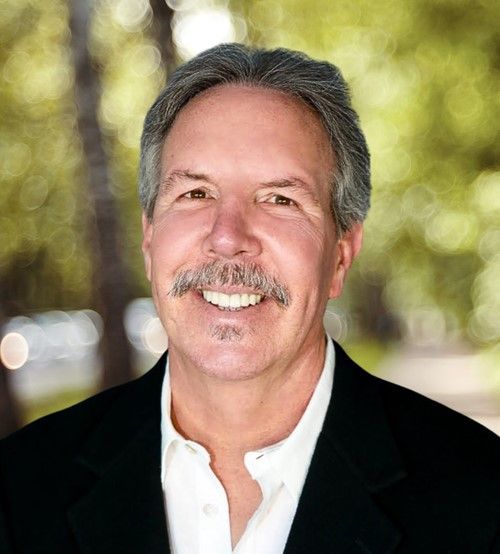 A man with a mustache smiling in a black blazer and white shirt against a soft-focus, leafy green background.