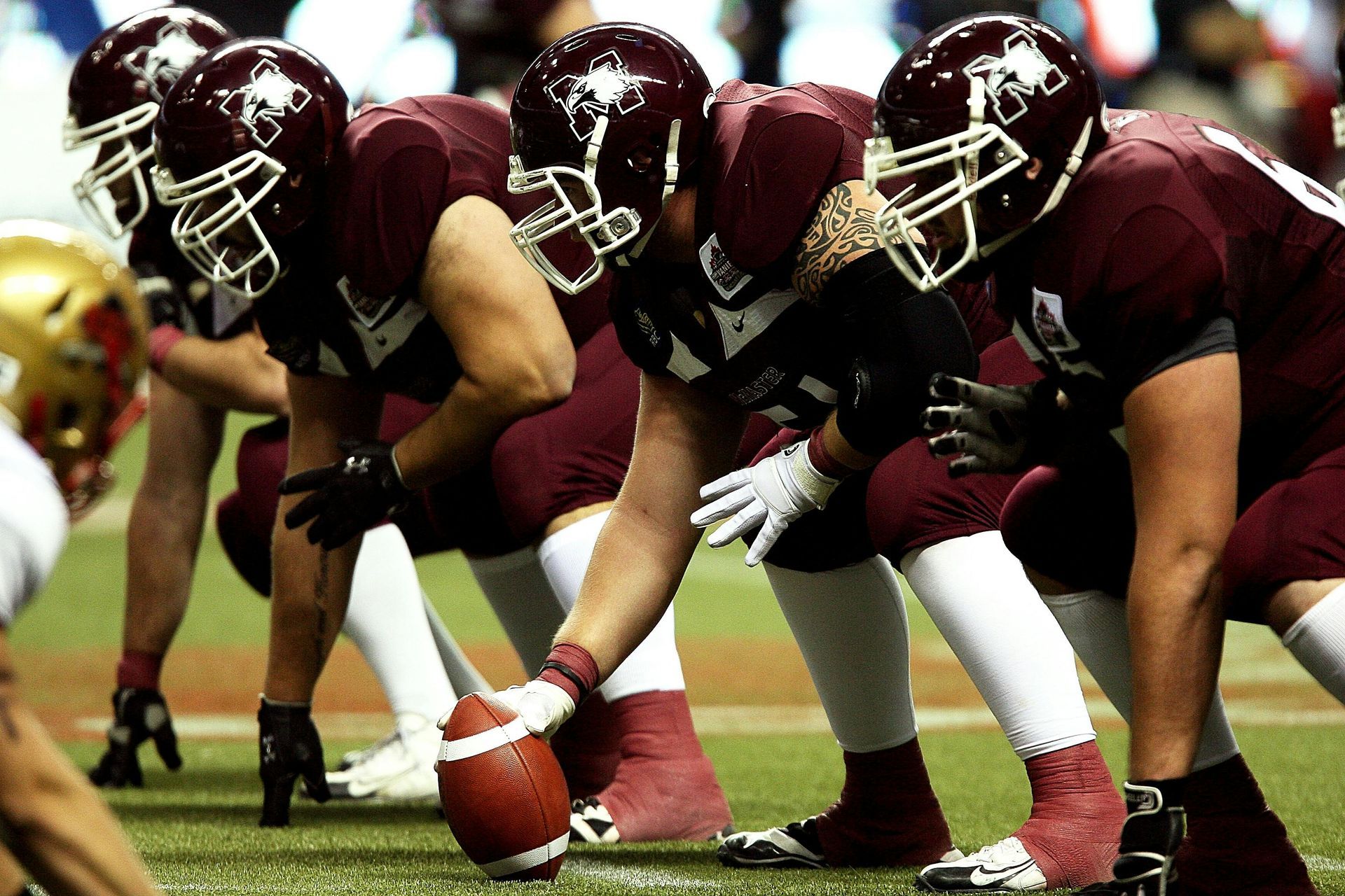 Football players in maroon helmets and jerseys lined up on the field, focused on the ball.