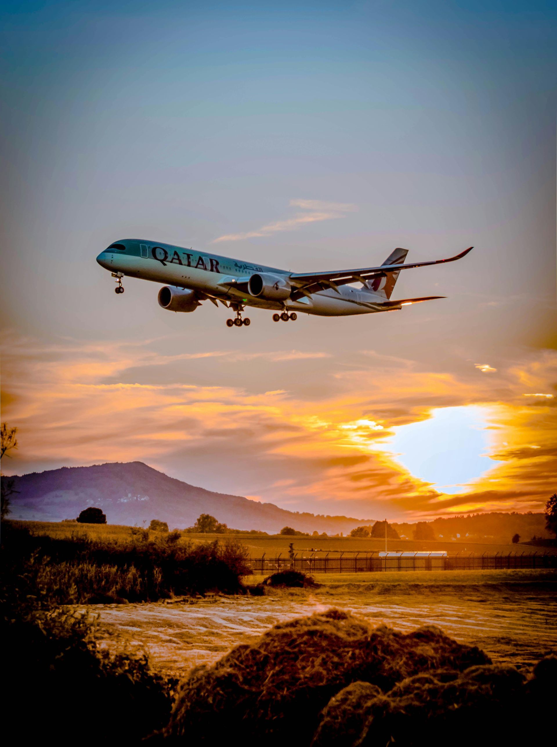 Qatar Airways airplane landing at sunset, silhouetted against a golden sky and landscape.