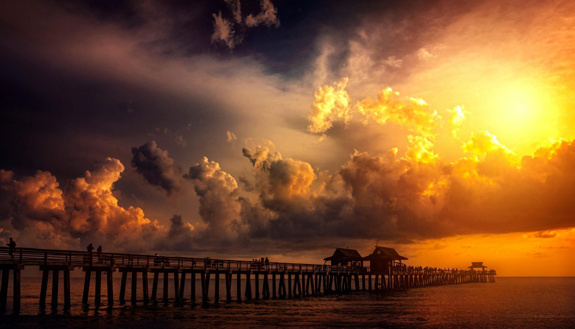Wooden pier extends into ocean at sunset, dramatic sky with sun bursting through clouds.