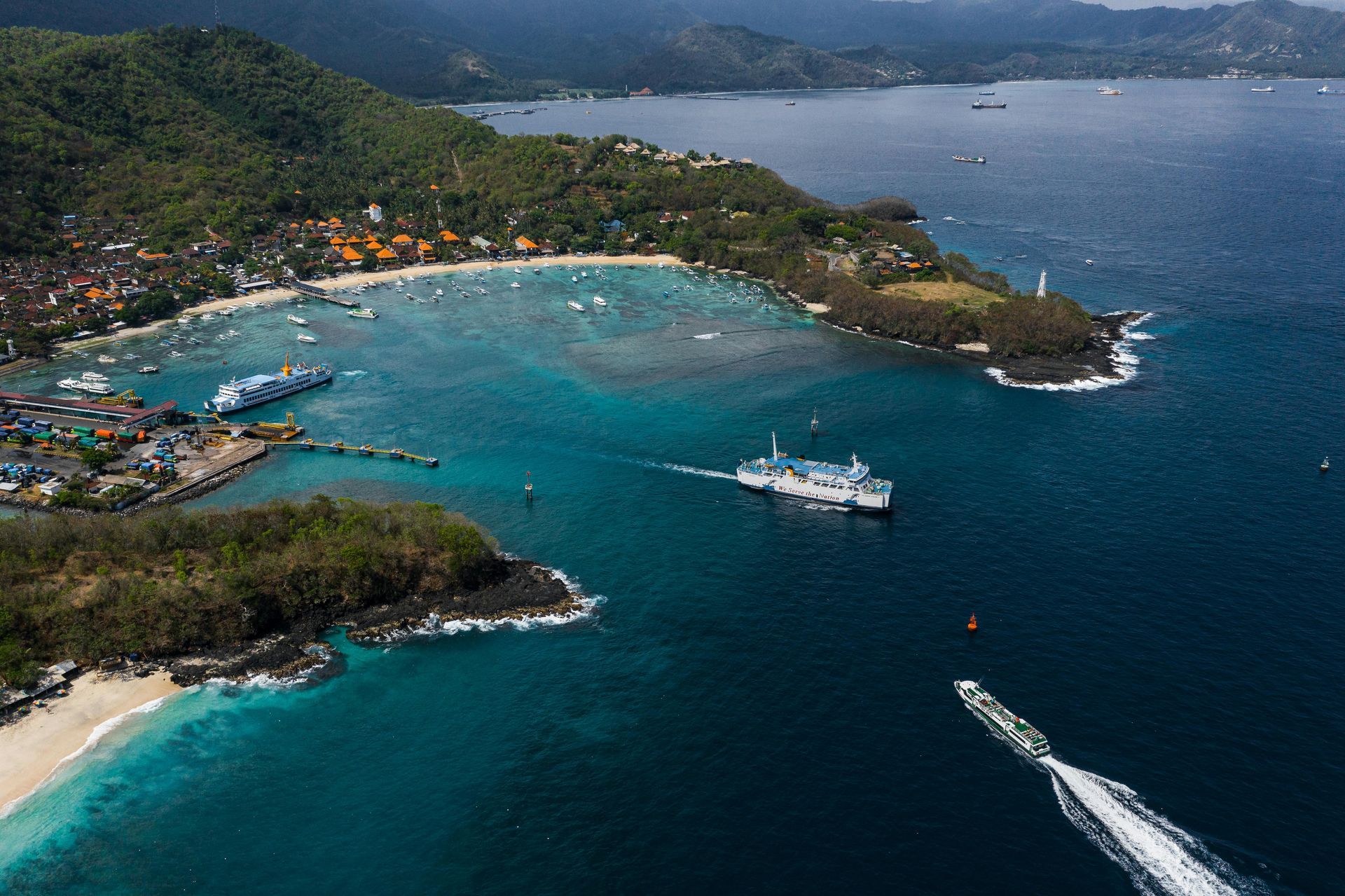 Aerial view of a coastal bay with turquoise water, boats, beaches, and a green, hilly shoreline.