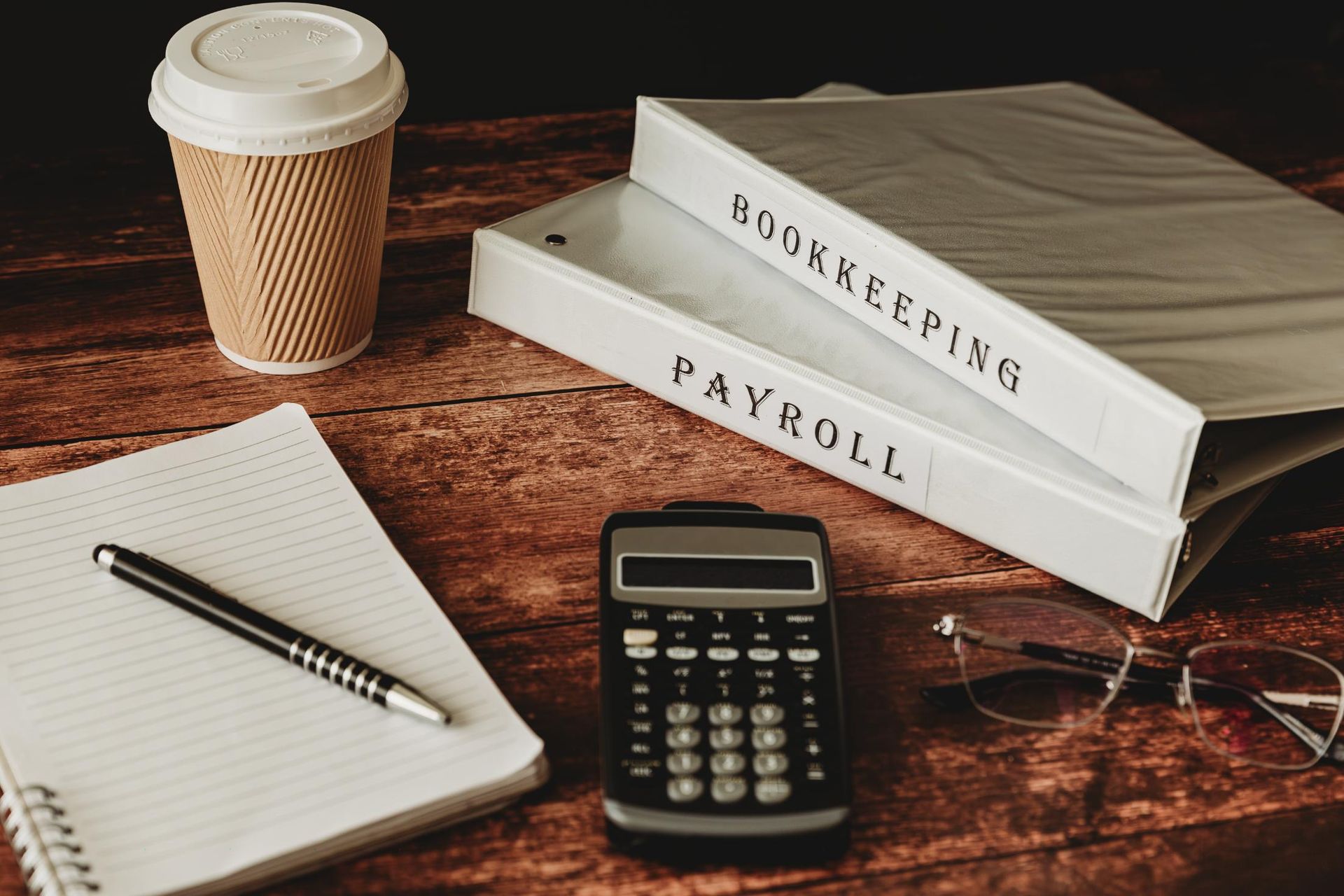 A calculator, notebook, pen, glasses and a cup of coffee are on a wooden table.