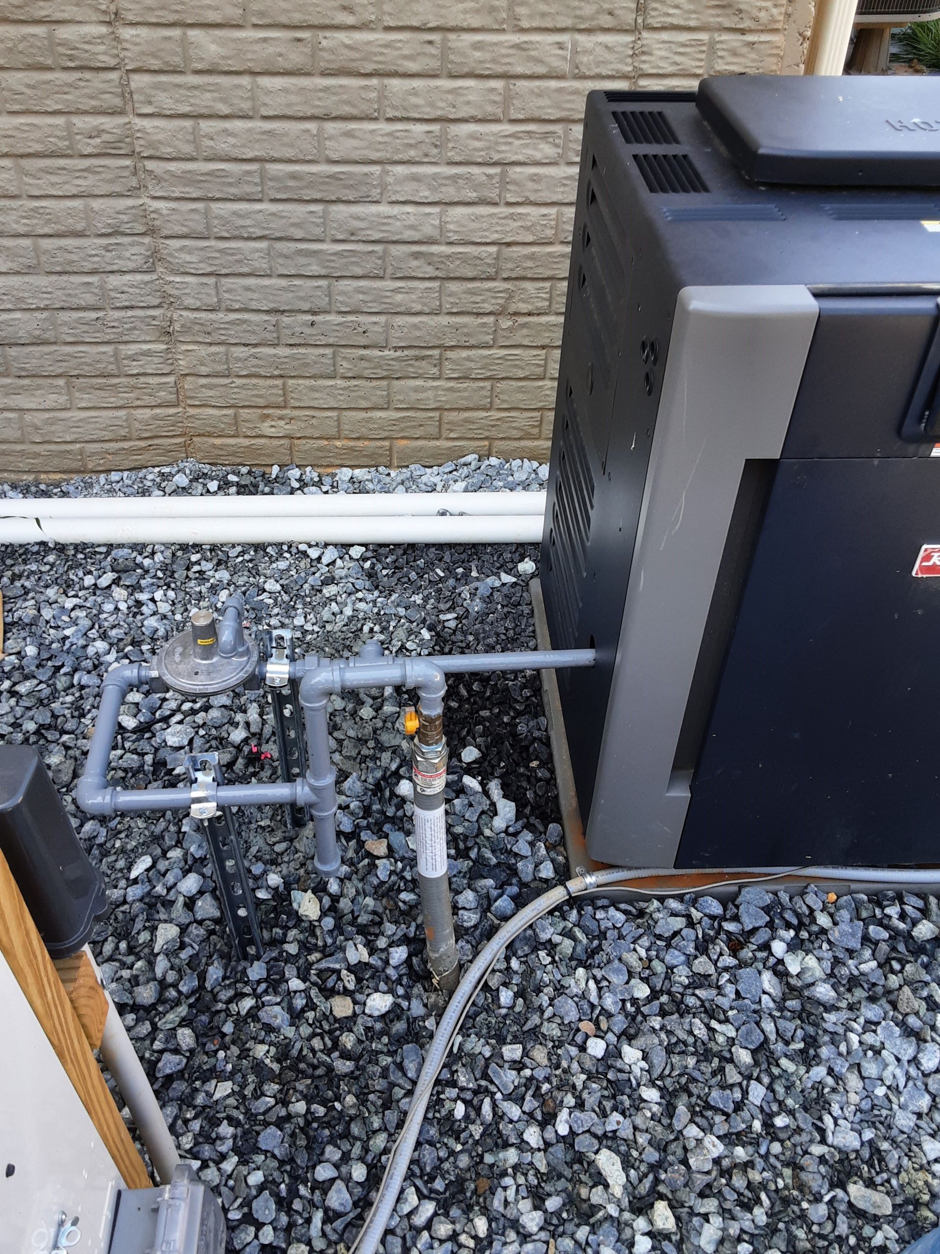 Pool heater with plumbing against a stone wall and gravel ground.