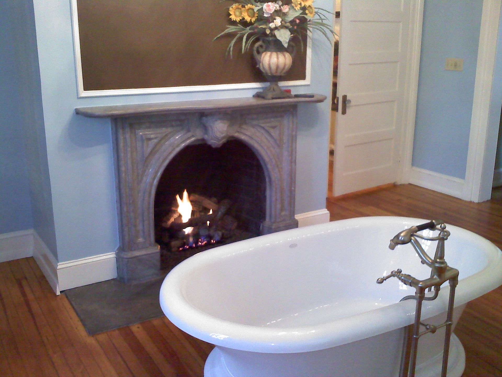 Bathroom with a bathtub in front of a fireplace with a lit fire; blue walls and wood floors.