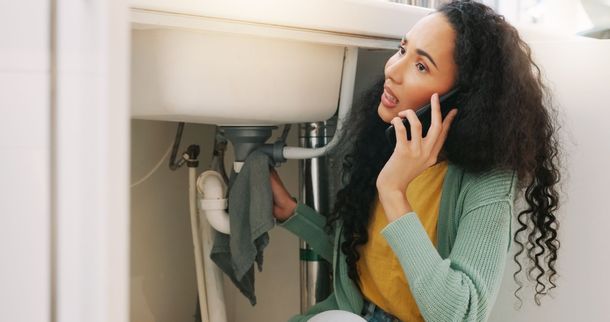 A woman is sitting under a sink talking on a cell phone.