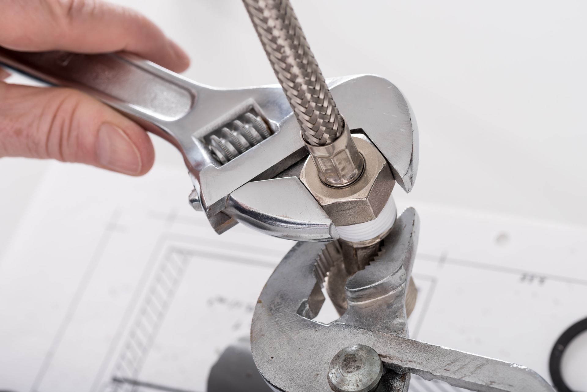 A hand uses an adjustable wrench and pliers to tighten a metal hose fitting over a technical blueprint. A hand uses an adjustable wrench and pliers to tighten a metal hose fitting over a technical blueprint.