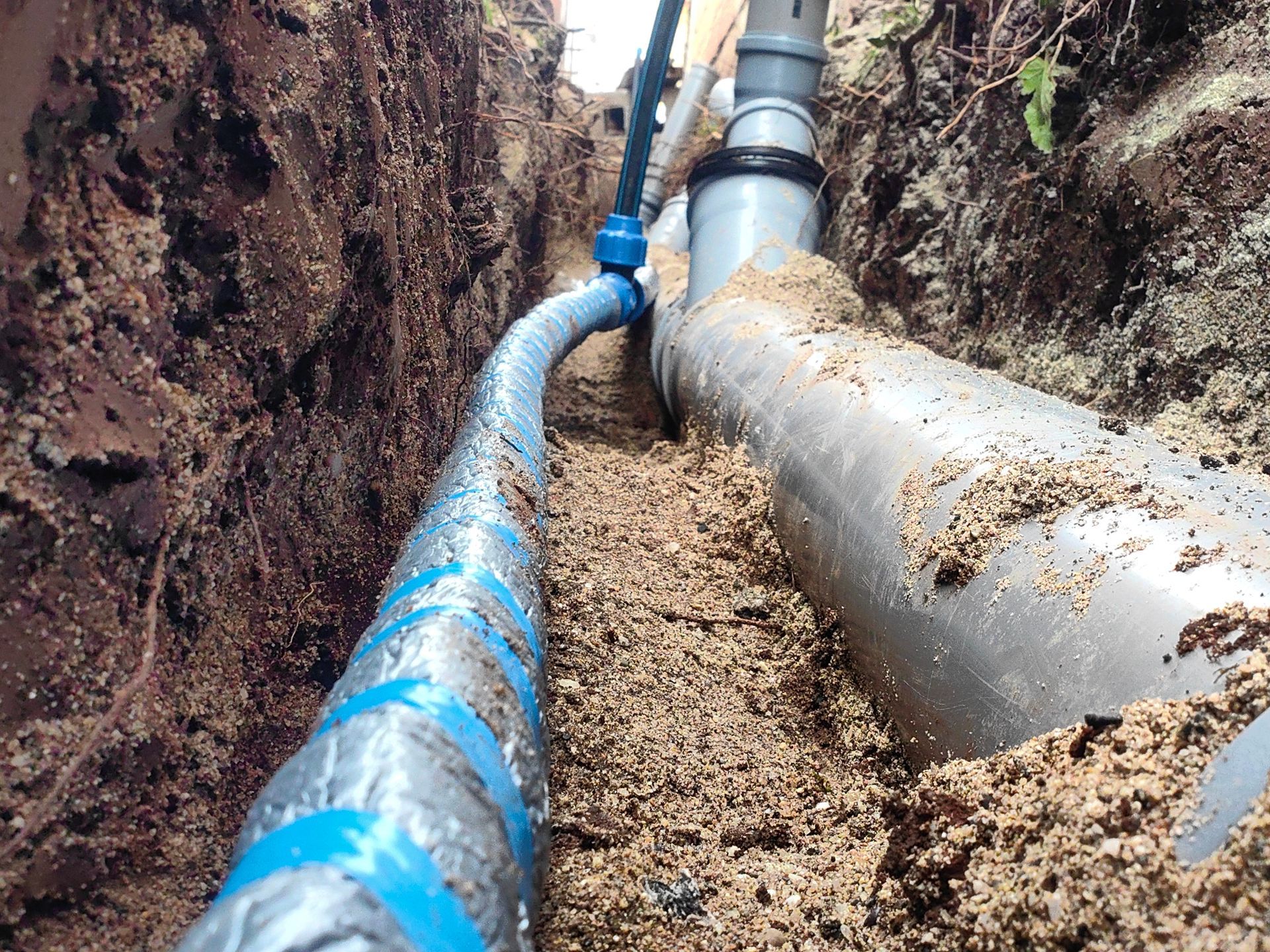 A trench shows a blue polyethylene water pipe running alongside a larger grey PVC drainage pipe in a sandy bed.