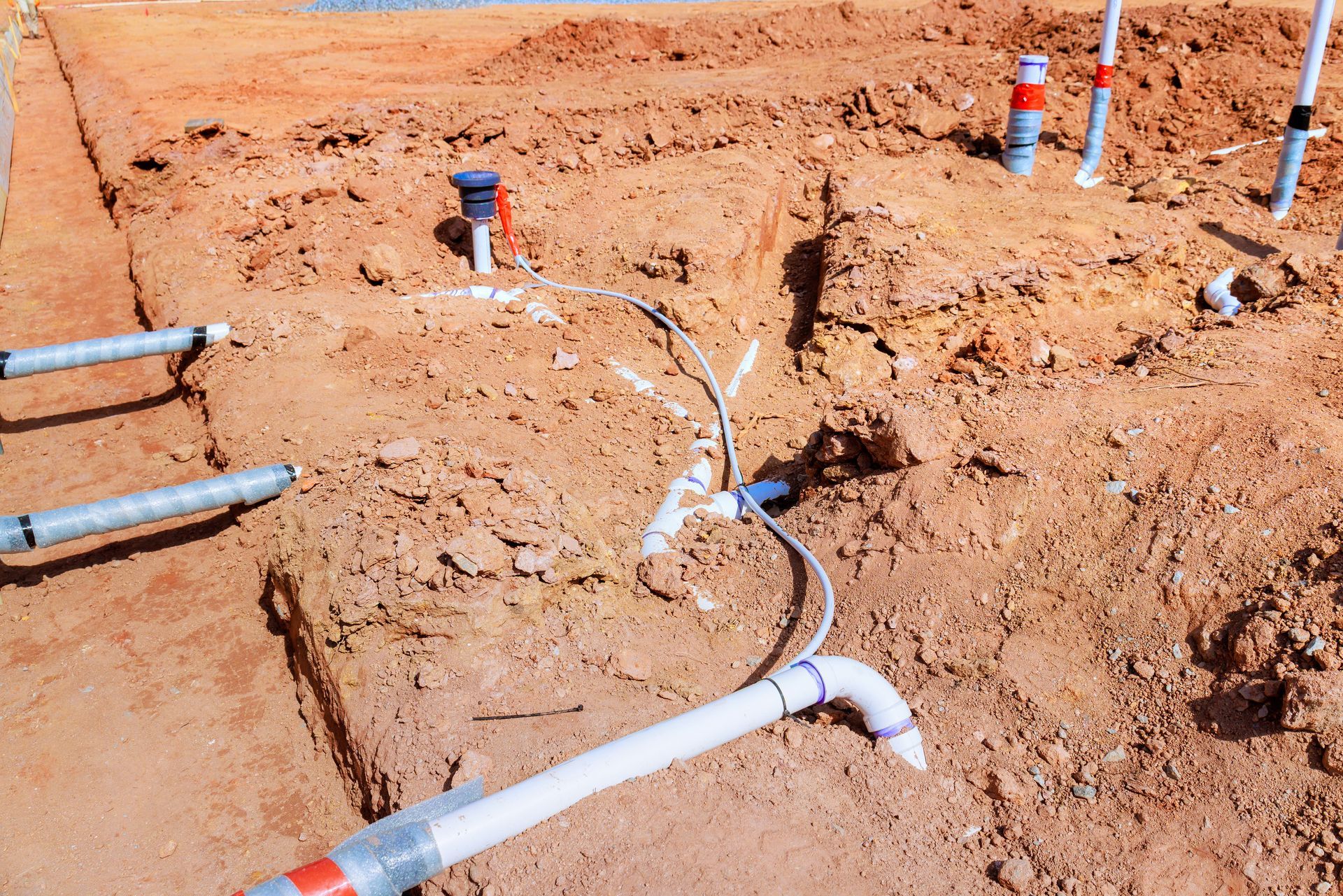 Construction site showing white PVC pipes, electrical conduits, and wiring laid out in dirt trenches for slab foundation.
