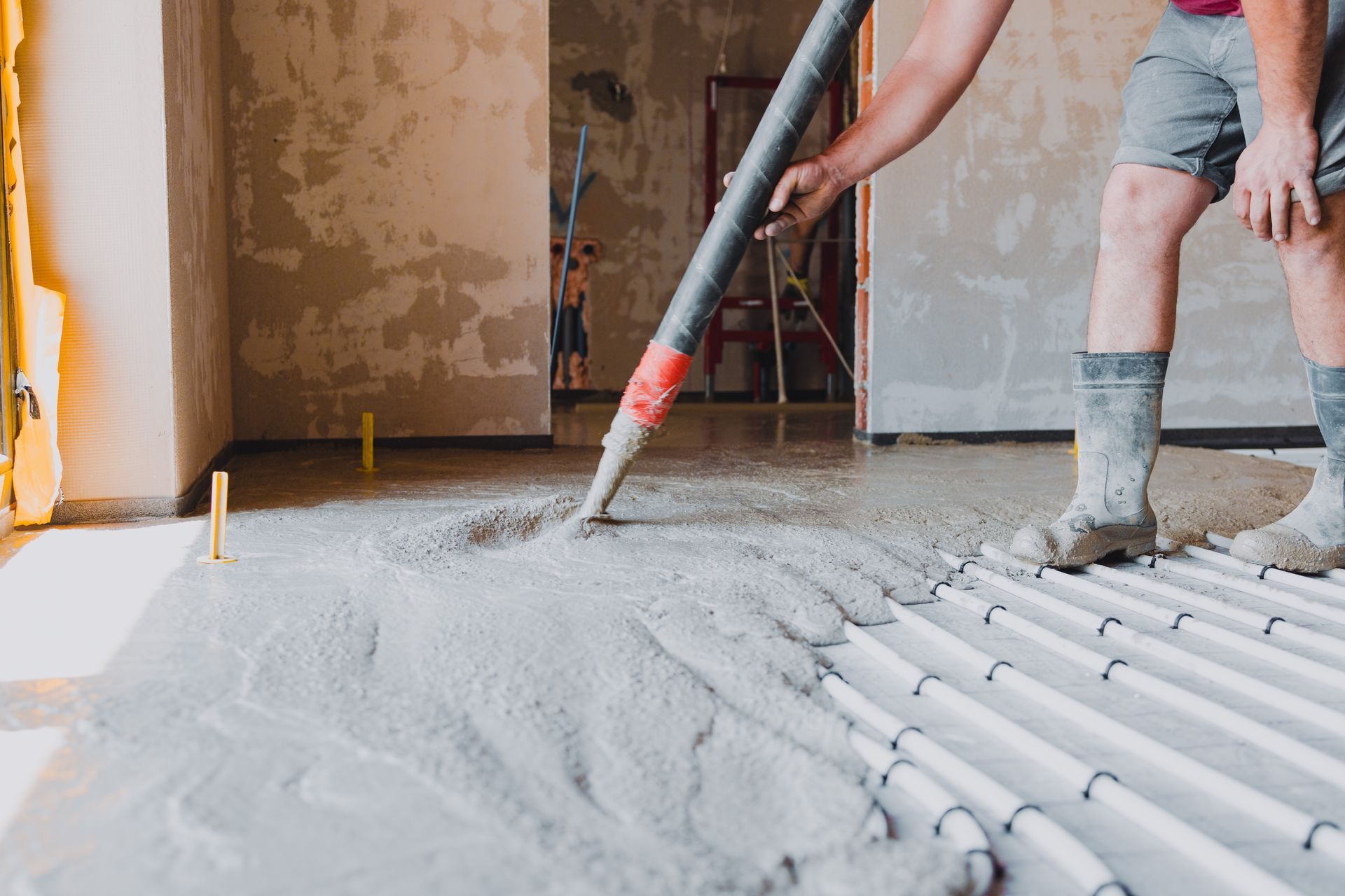 A worker in boots uses a hose to pour liquid concrete over radiant floor heating pipes in a room under construction.
