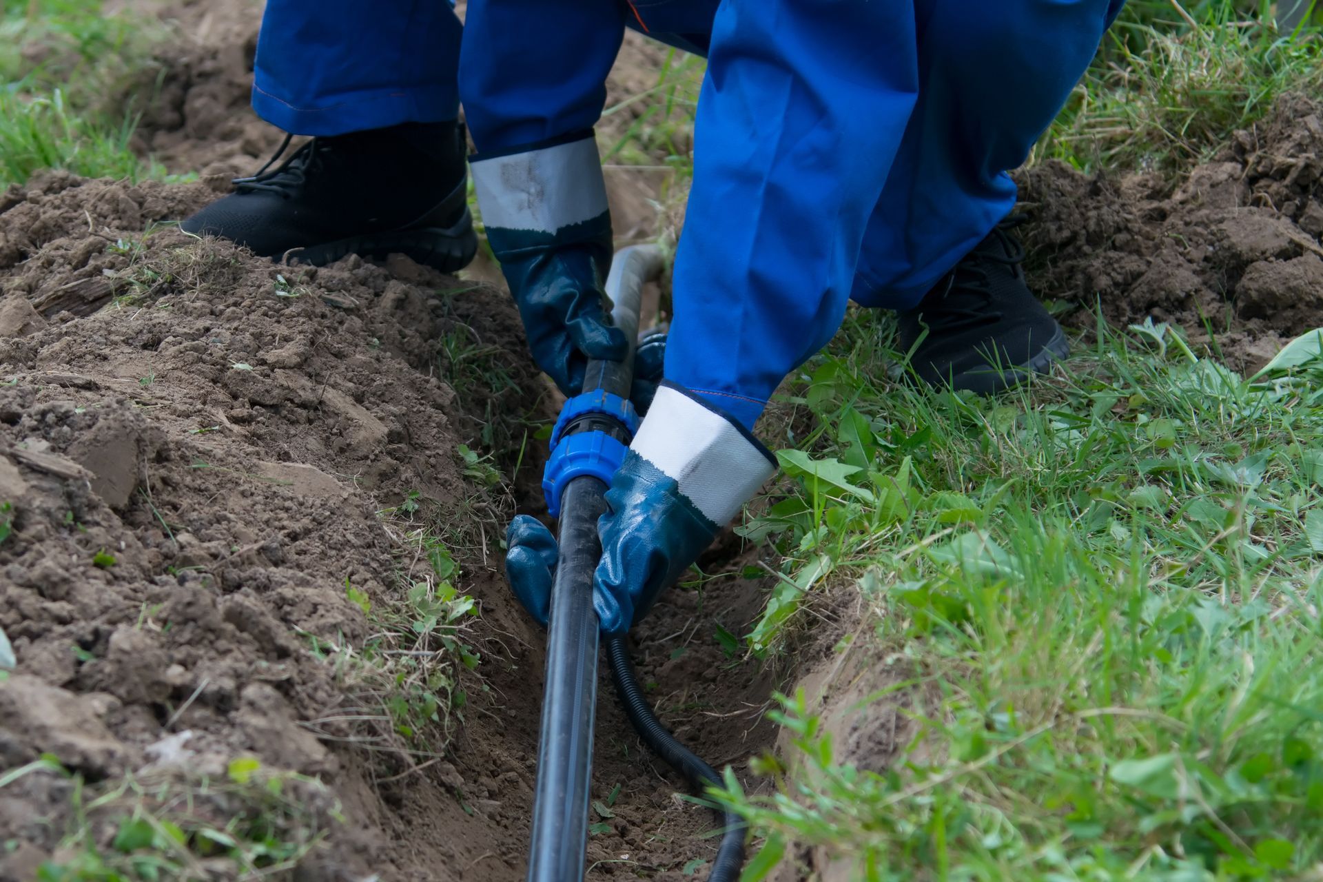 A person in blue coveralls and gloves installs a black pipe in a shallow trench in the ground.